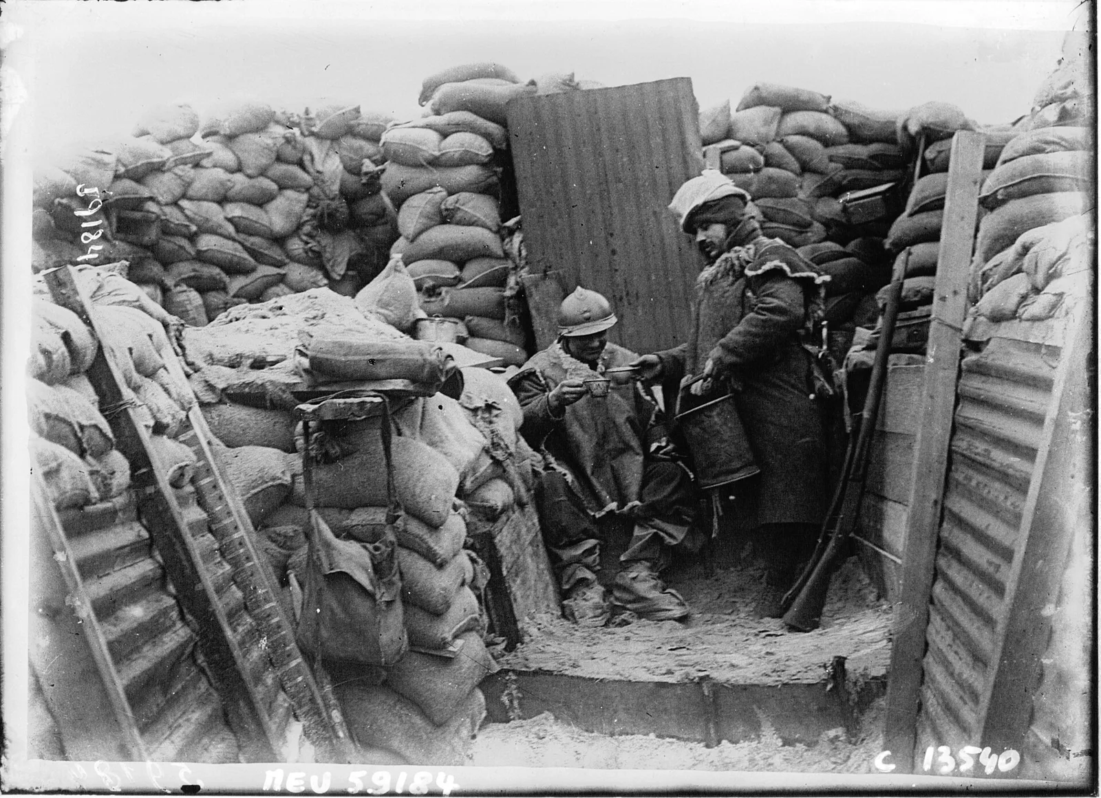 Des soldats français dans une tranchée de première ligne, entre des sacs de terre, en janvier 1916 : l’un tend son quart, l’autre lui verse le café.